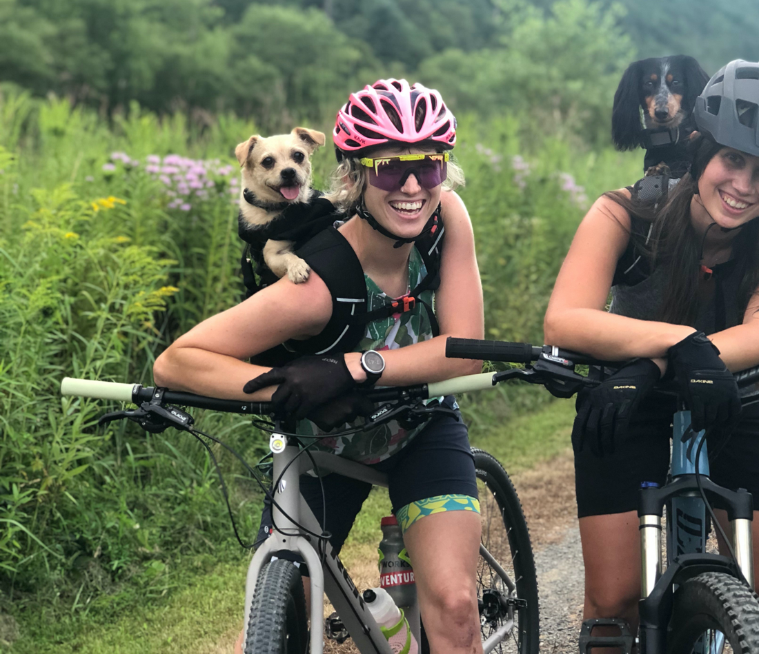 Two ladies resting on their bikes with their backpack carriers lifting their dogs.