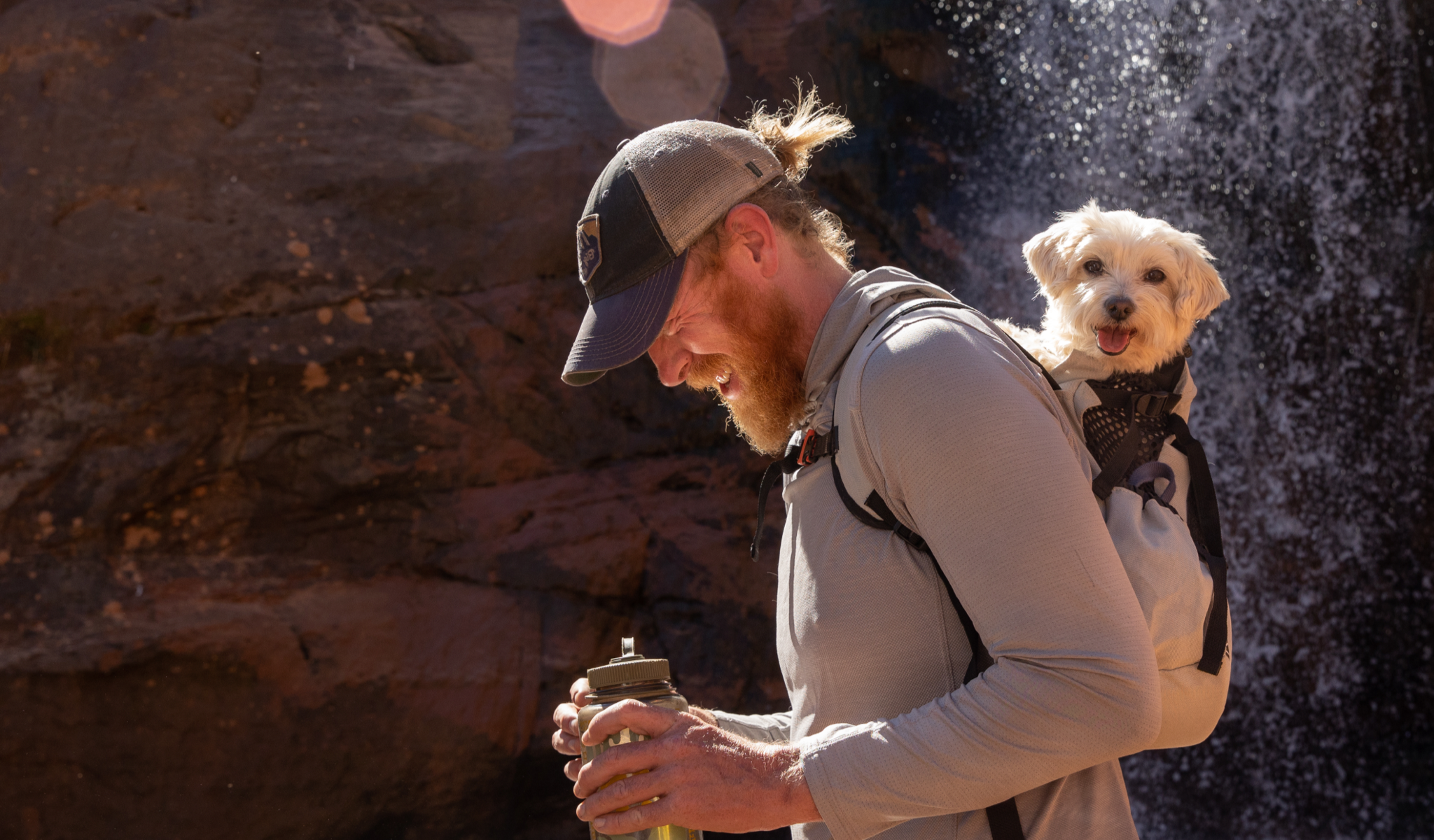 A man wears his dog on his back in an AIR2 backpack next to a waterfall.