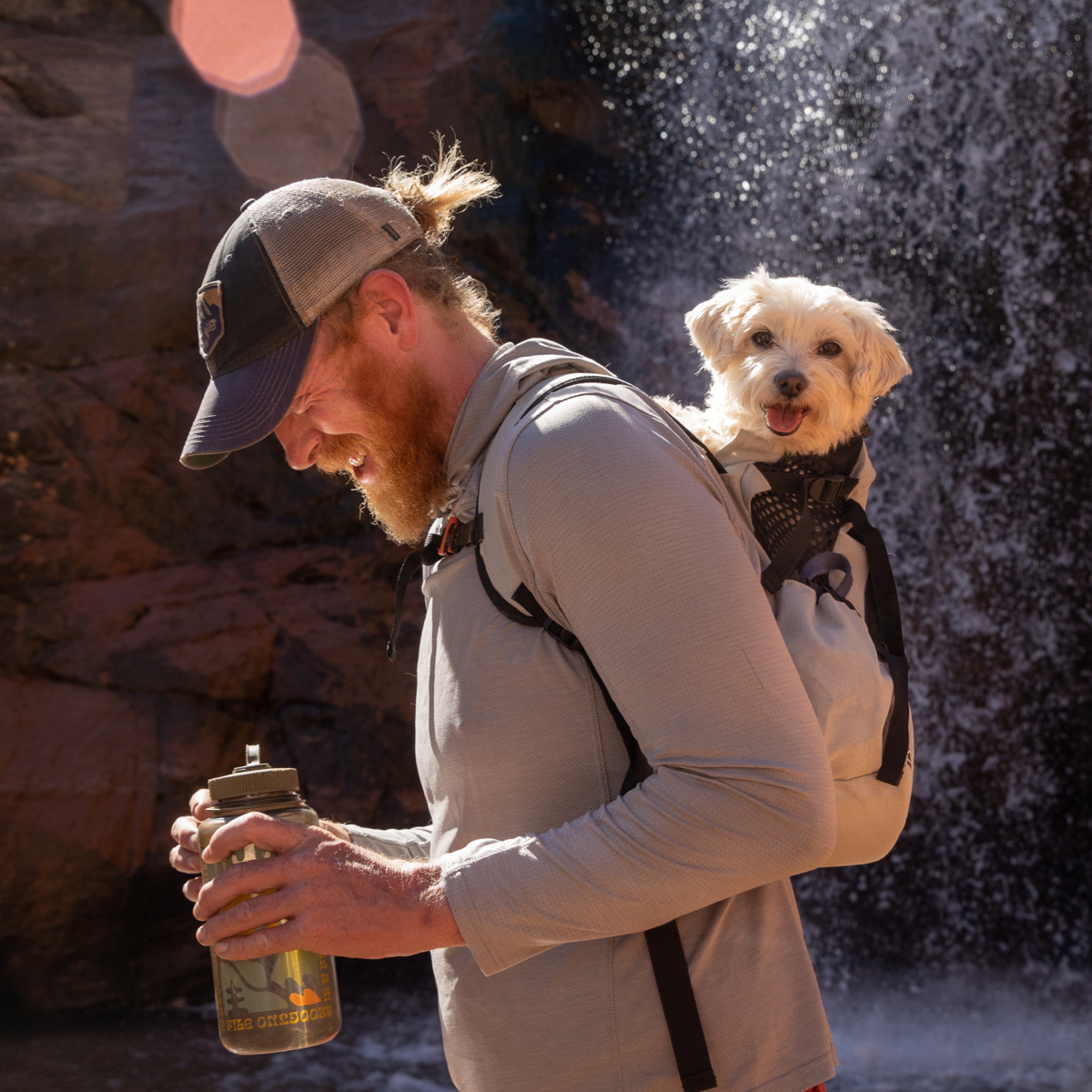 A man wears his dog on his back in an AIR2 backpack next to a waterfall.