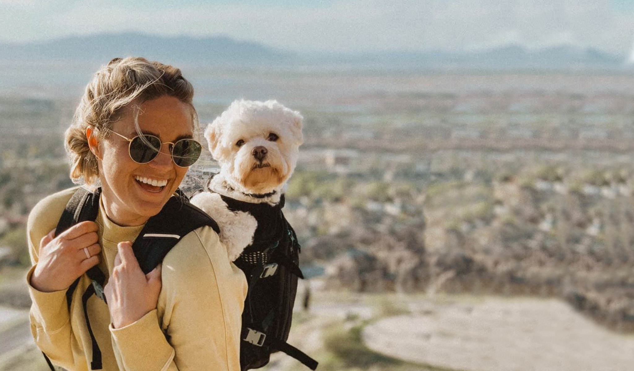 A woman wearing sunglasses and overlooking a hillside with a white bichon frise in her black K9 Sport Sack Plus 2 on her back