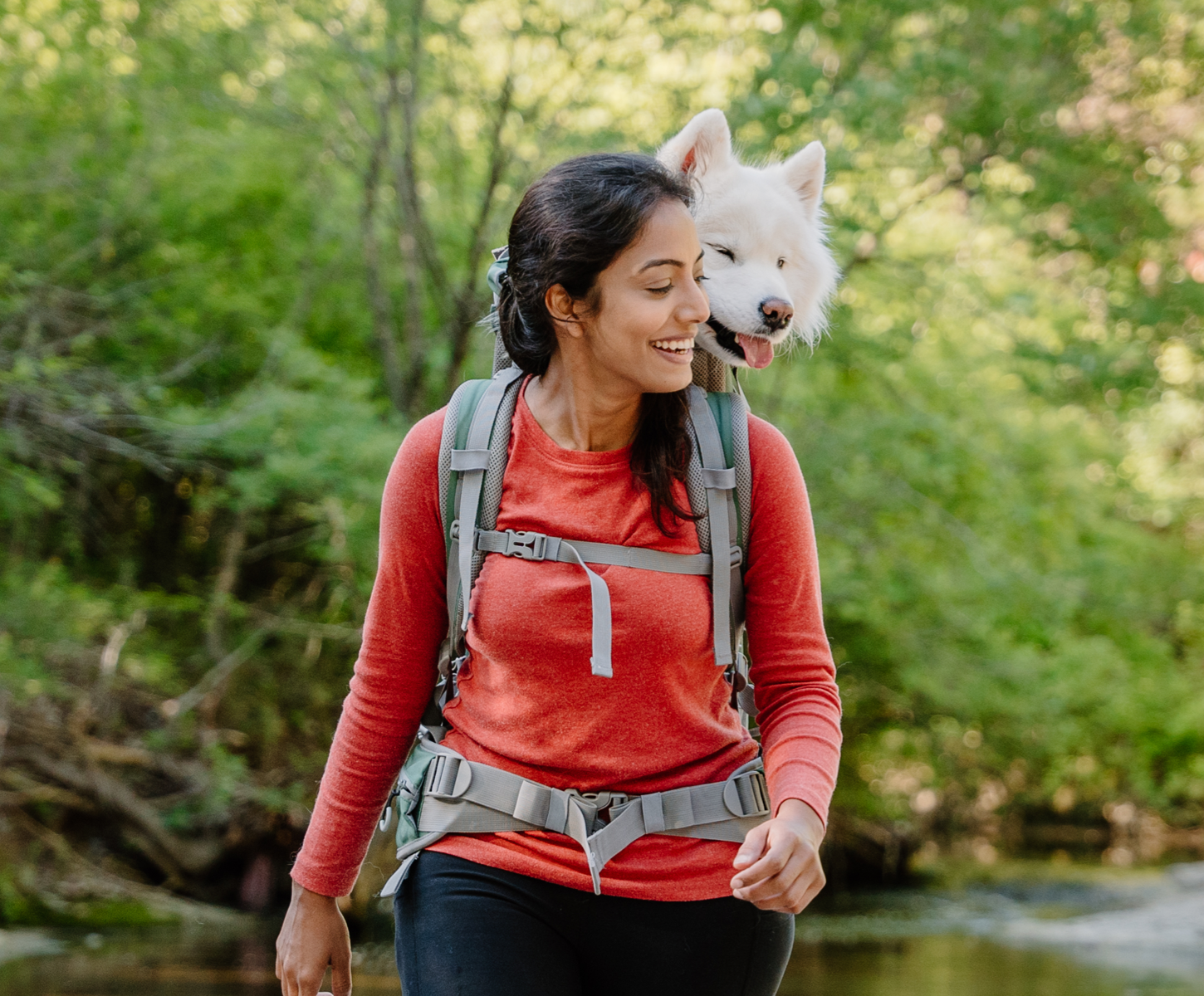 A youthful woman and her white husky stroll through the woods using a green K9 Sport Sack Rover 2 backpack carrier