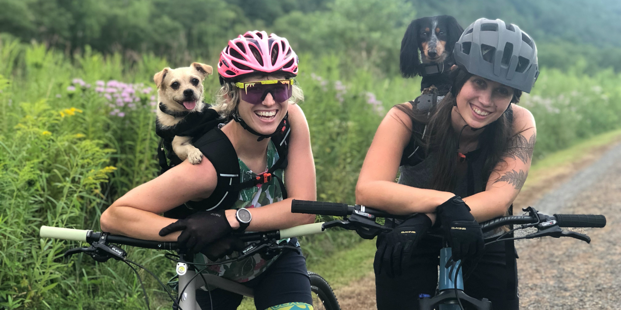 Two ladies resting on their bikes with their backpack carriers lifting their dogs.